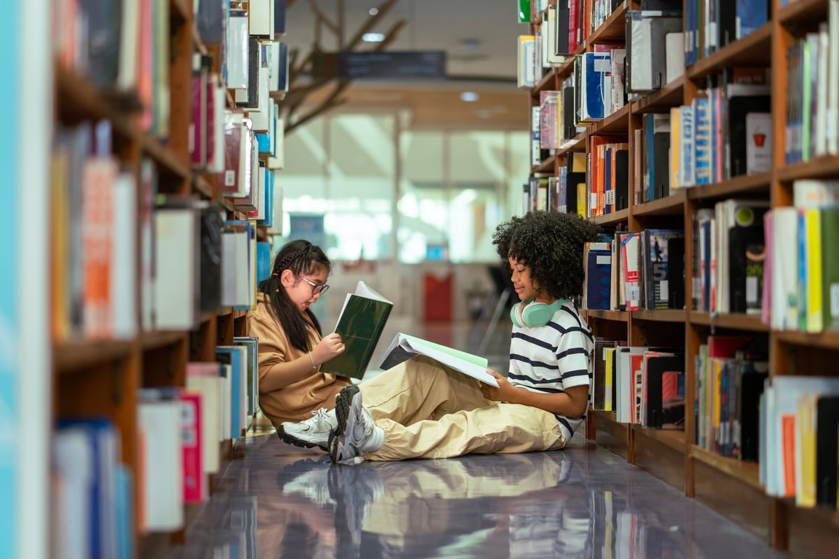 Student girls sitting on the floor reading book between bookshelf in school library