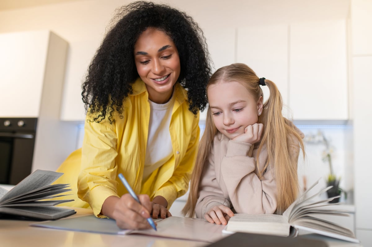 Curly woman writing in notebook near girl at home during tutoring session