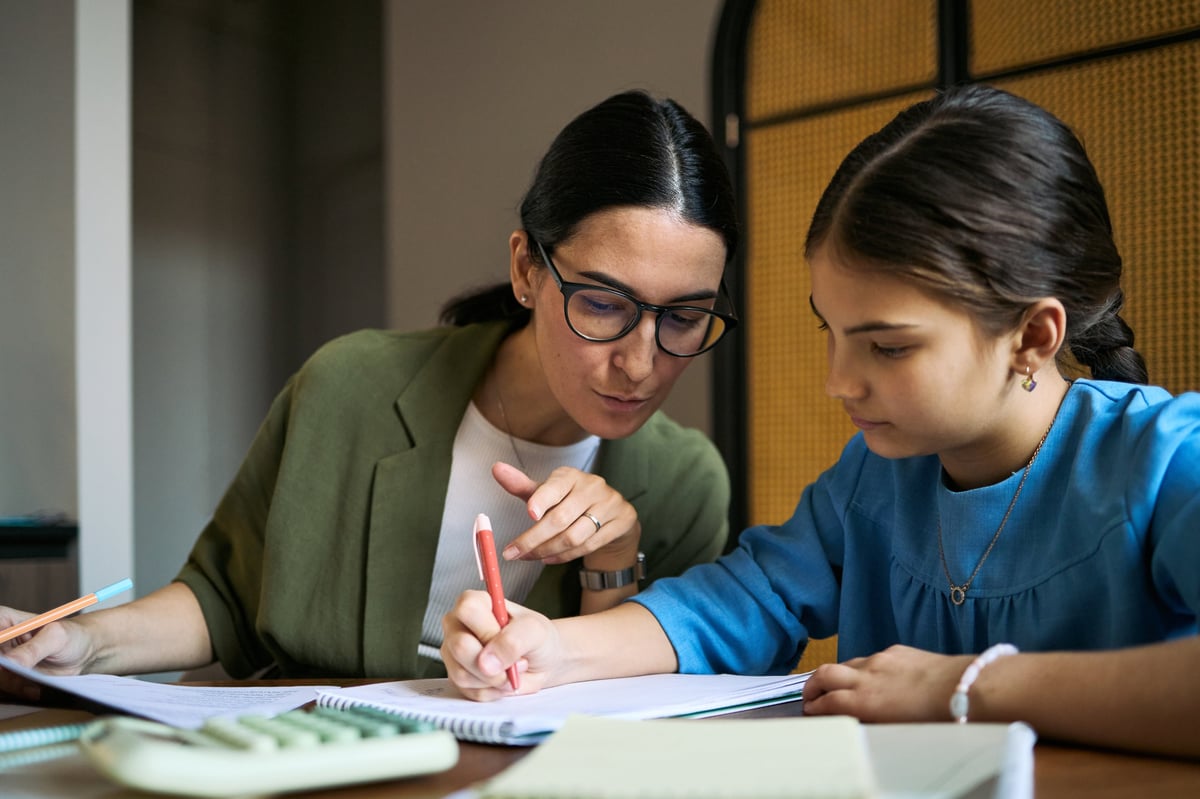 Middle aged woman guiding schoolgirl while writing in workbook during tutoring session