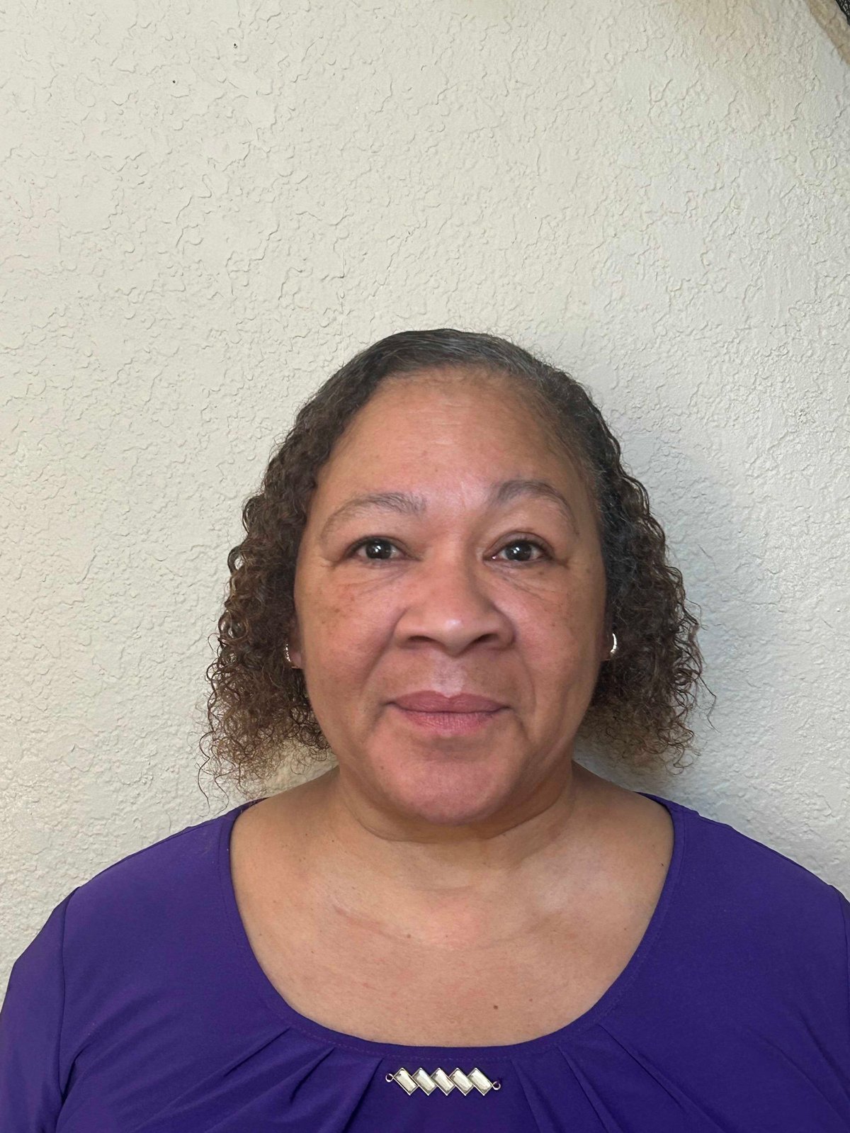 Portrait of a middle-aged woman with curly brown hair wearing a purple top against a white textured wall