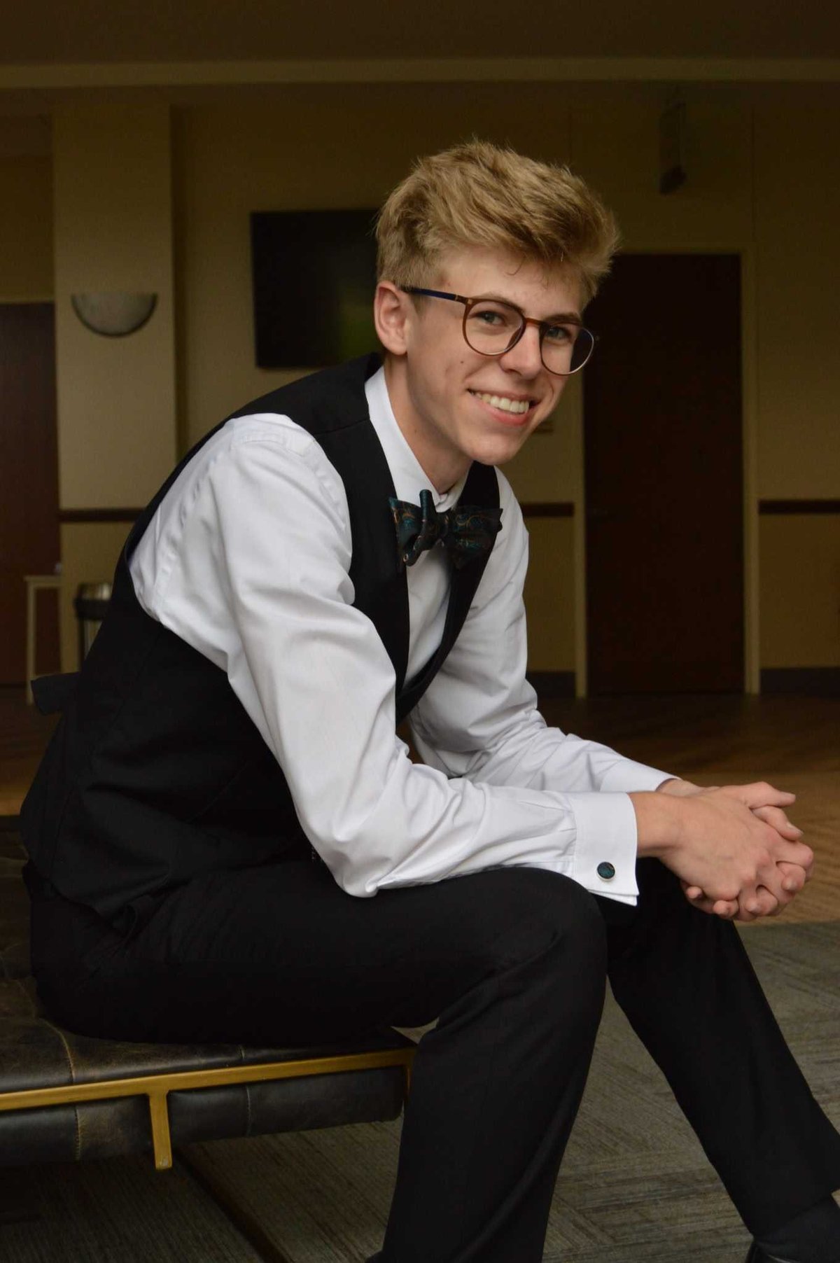 Young man in formal attire wearing glasses, white dress shirt, black vest and bow tie, smiling while seated indoors
