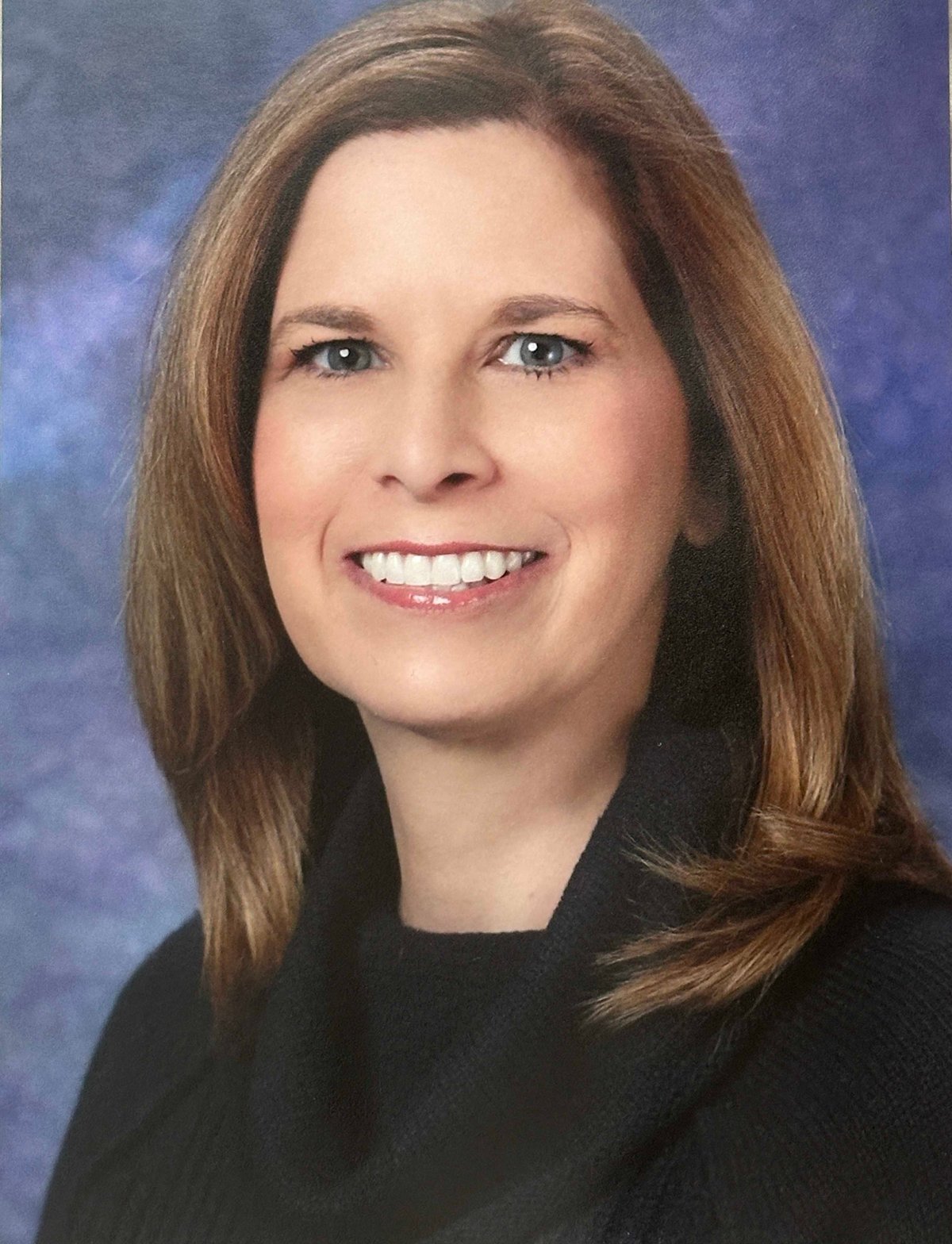 Professional headshot of a smiling woman with reddish-brown hair wearing a black blazer against a blue background