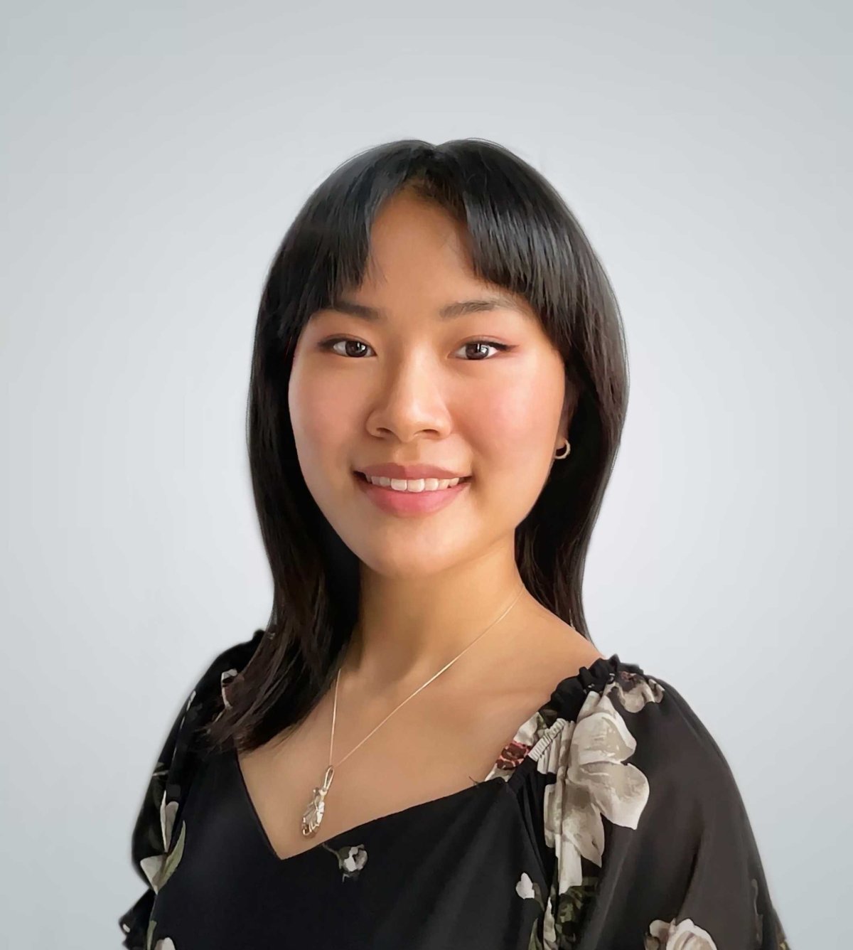 Portrait of a young woman with long black hair, bangs, and a warm smile wearing a black floral top with a pendant necklace