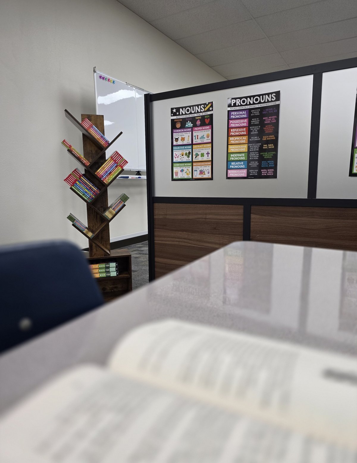 Classroom corner with grammar posters on wall, wooden tree-shaped book shelf with colorful books, and white table in foreground